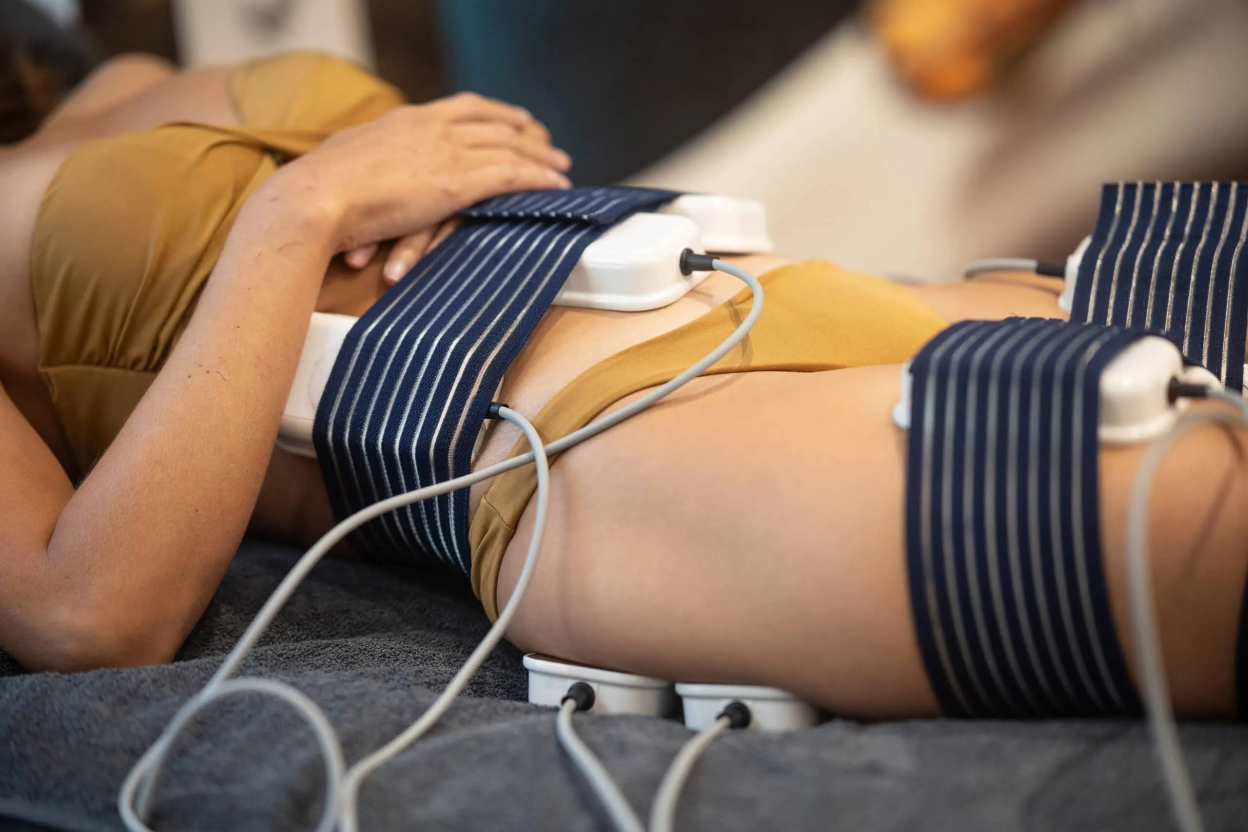 Das Bild zeigt eine Frau in entspannter Rückenlage, deren Bauch- und Oberschenkelbereich mit EMS-Pads zur schmerzfreien Muskelstimulation und Behandlung umwickelt ist.